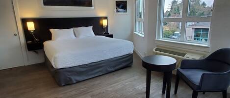 A hotel room with a large bed, two bedside tables with lamps, a small round table, a chair, and a window with a view of trees and buildings.
