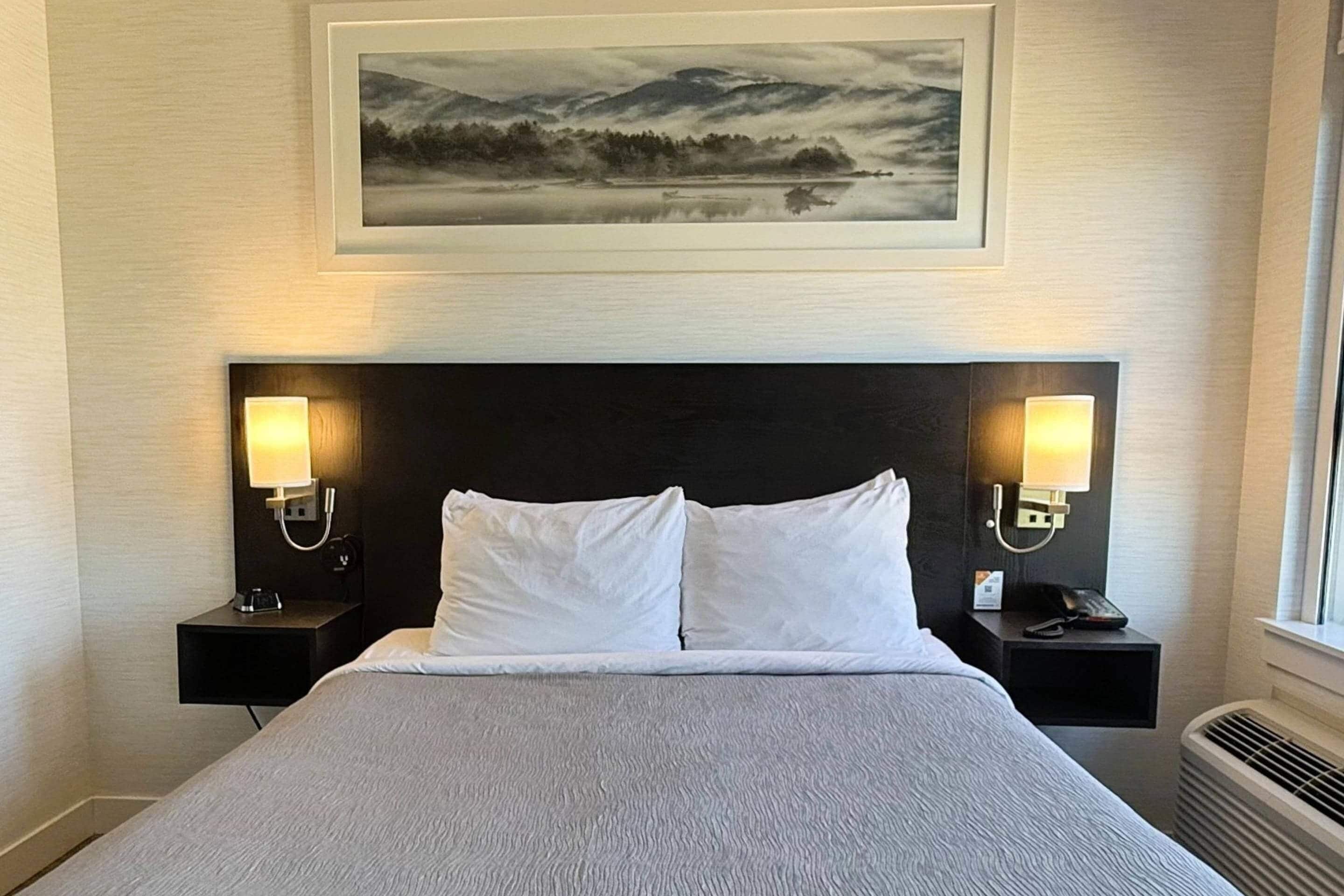 A hotel room with a large bed, two bedside tables with lamps, a framed picture above the bed, and a window on the right.