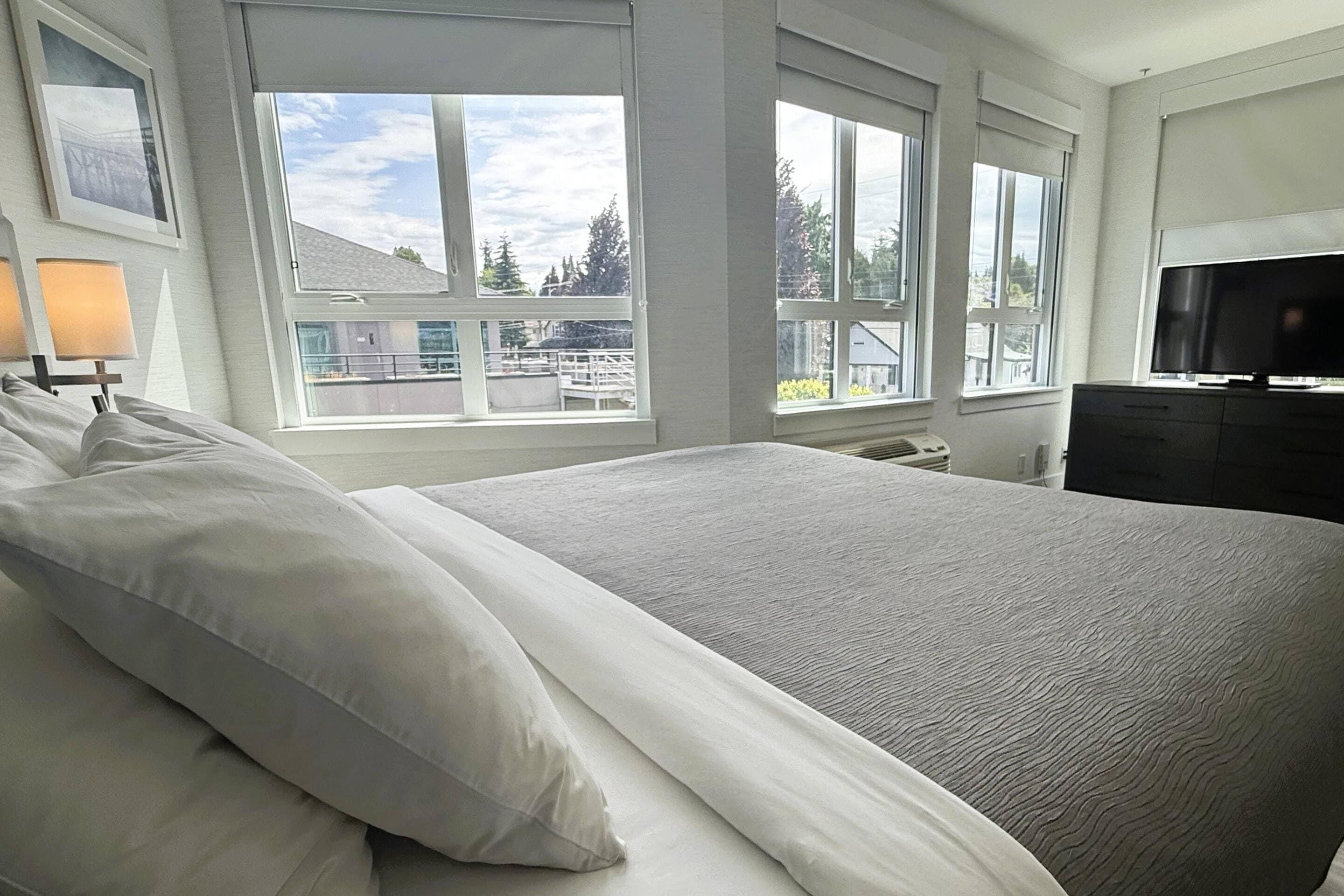 A large bed with white linens, a television on a stand, and large windows with a view of buildings and trees.