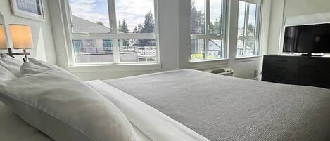 A large bed with white linens, a television on a stand, and large windows with a view of buildings and trees.