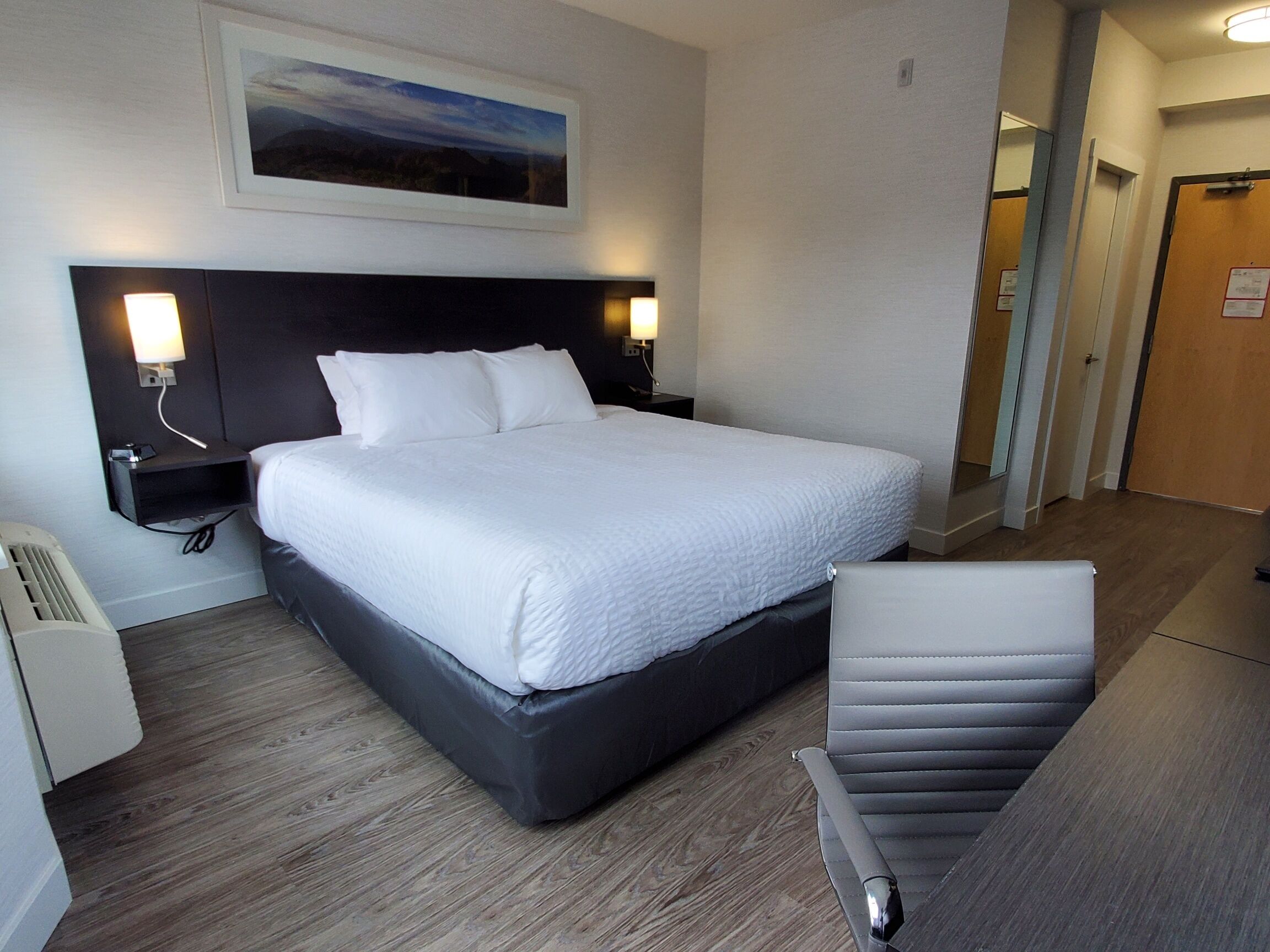 A hotel room with a large bed, bedside lamps, a chair, a desk, and a framed picture on the wall.