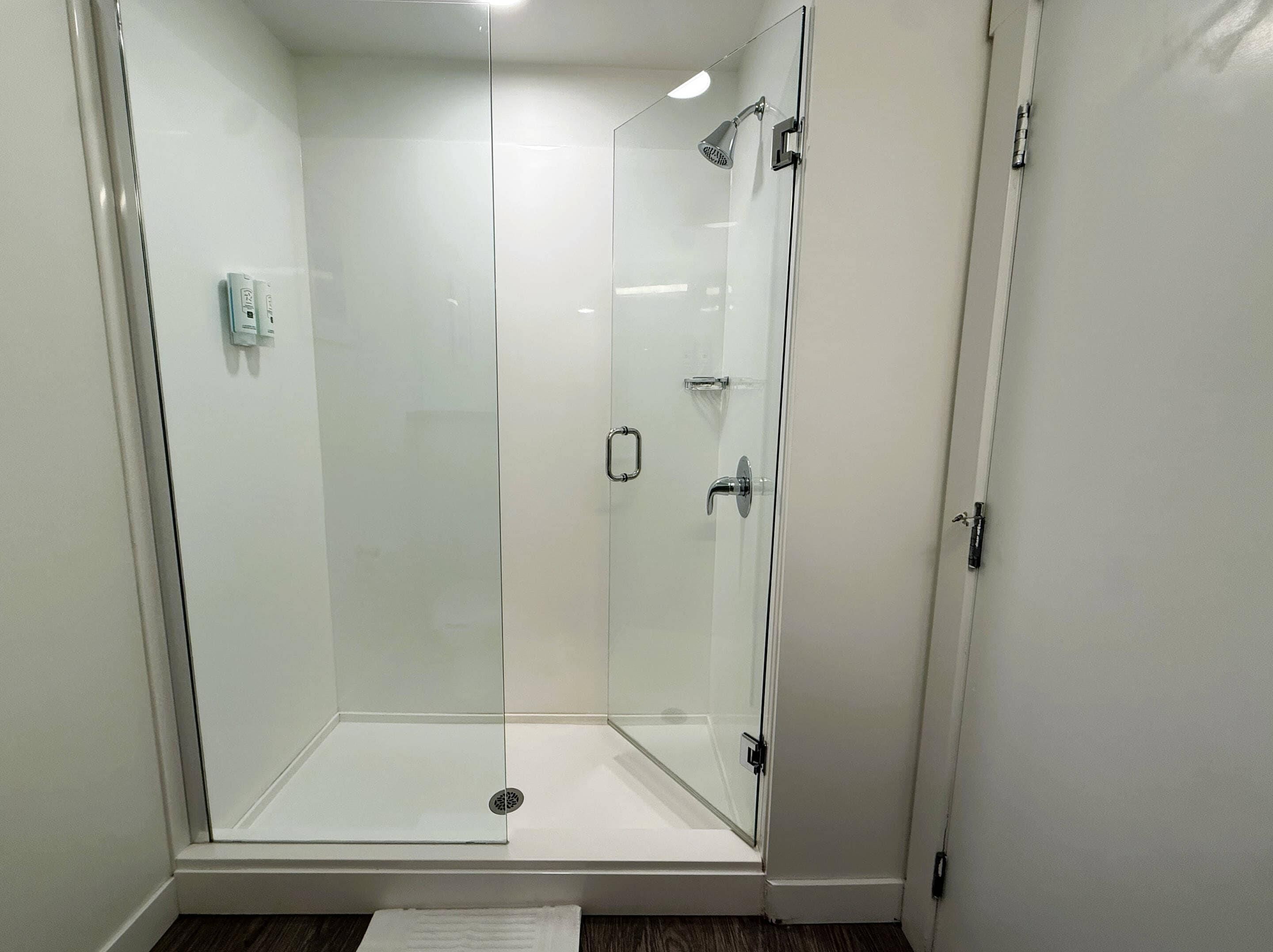Une douche fermée par des parois en verre, avec une porte blanche, un interrupteur visible et un drain situé en bas.