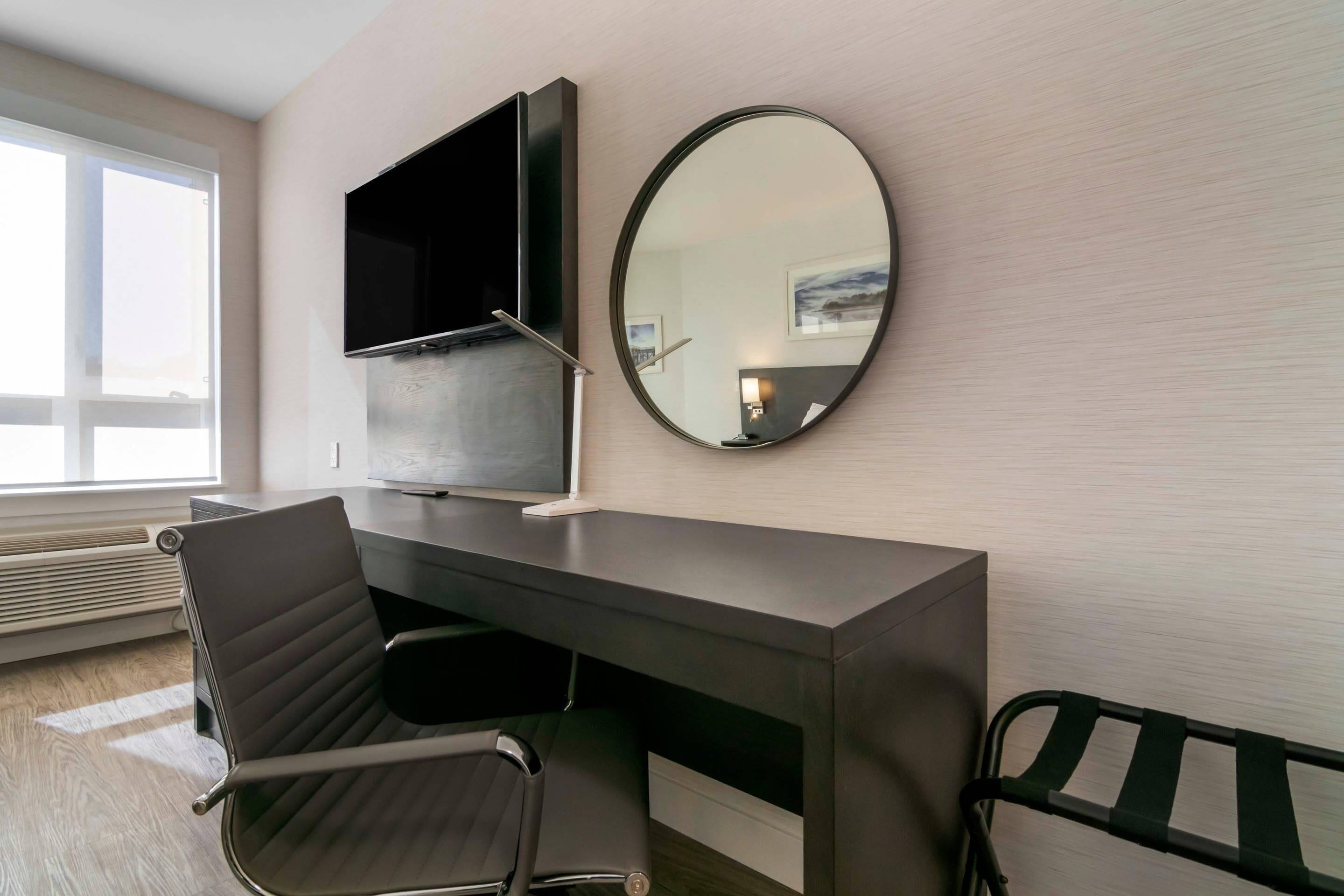 A modern hotel room with a desk, chair, TV, and a round mirror.