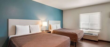 A hotel room with two beds, a window with blinds, a heater, and a nightstand with a lamp.