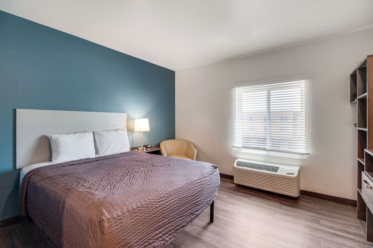 Une chambre d’hôtel avec un grand lit, une chaise, une table de chevet, une lampe, une fenêtre avec des stores et un système de climatisation intégré.