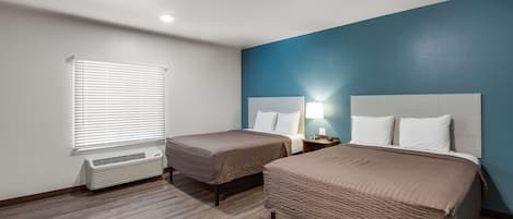 A hotel room with two beds, a window with blinds, an air conditioner, and a nightstand with a lamp.
