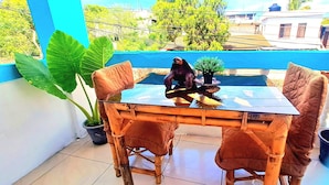 Une table à plateau en verre, avec une chaise, sur un balcon aux murs bleus, agrémenté de plantes en pot.
