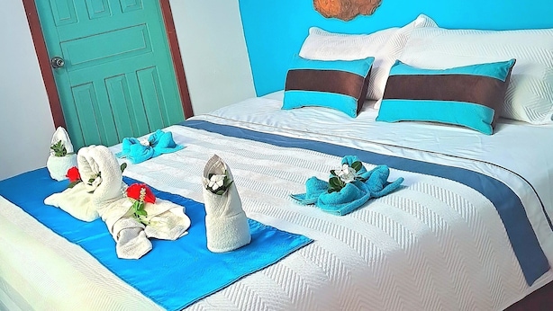 Une chambre d’hôtel avec un lit, un mur bleu orné d’œuvres d’art évoquant une île, une porte verte et un plafond blanc.