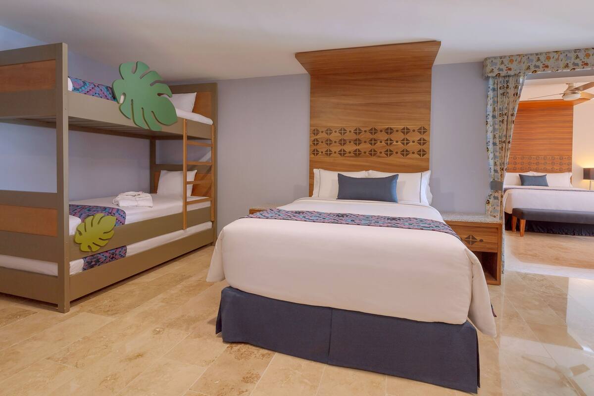 A hotel room with a bunk bed, a single bed, a wooden headboard, a nightstand, and a bedside table.