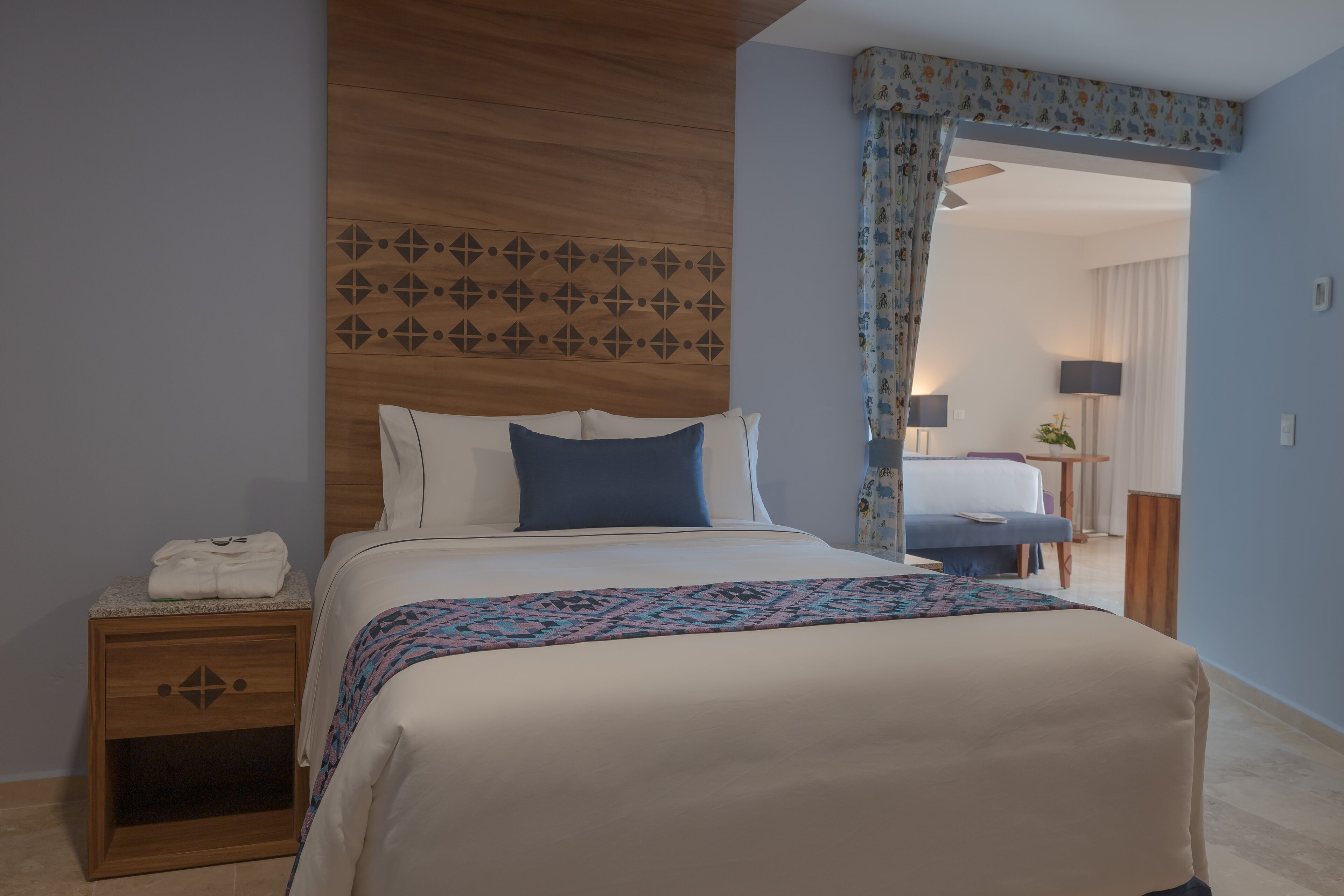 A hotel room with a bed, a nightstand, a patterned headboard, and a view of another room through a doorway.