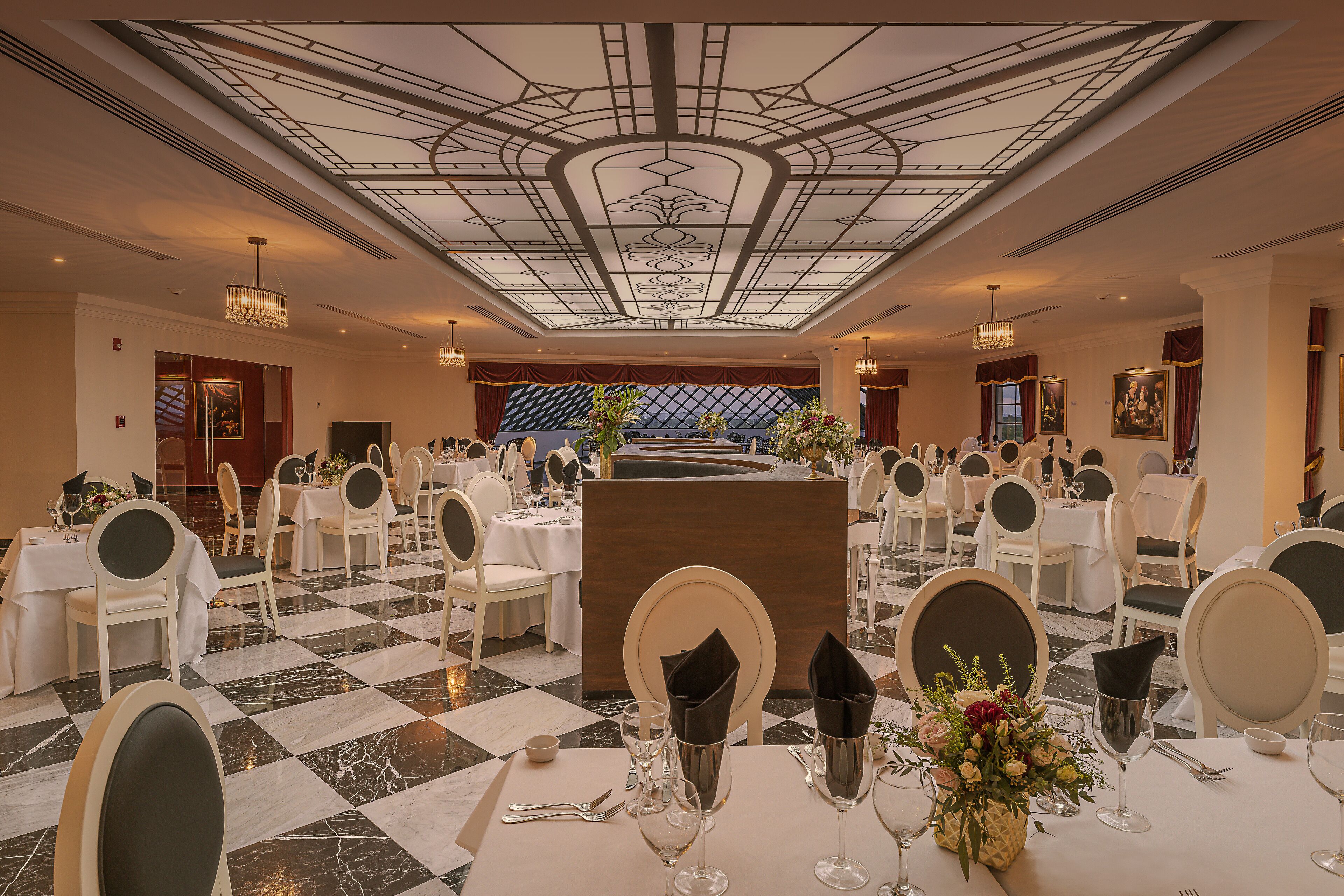 A formal dining room with a checkered floor, round tables, and chairs, a central podium, and a decorative ceiling.