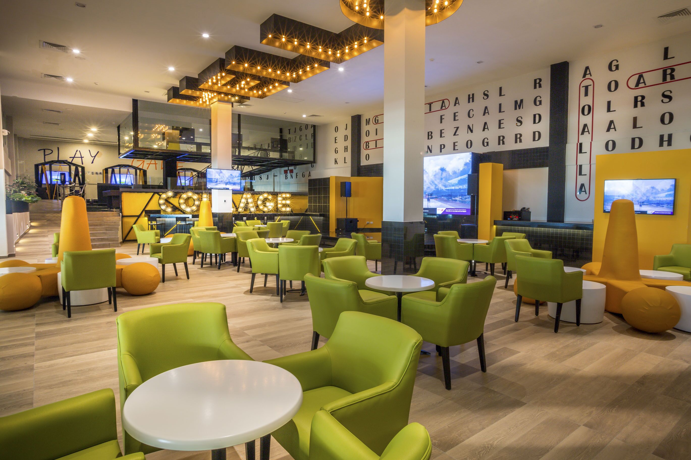 Un área de descanso moderna con asientos verdes y amarillos, una zona de bar con televisores y una escalera que conduce a un nivel superior.