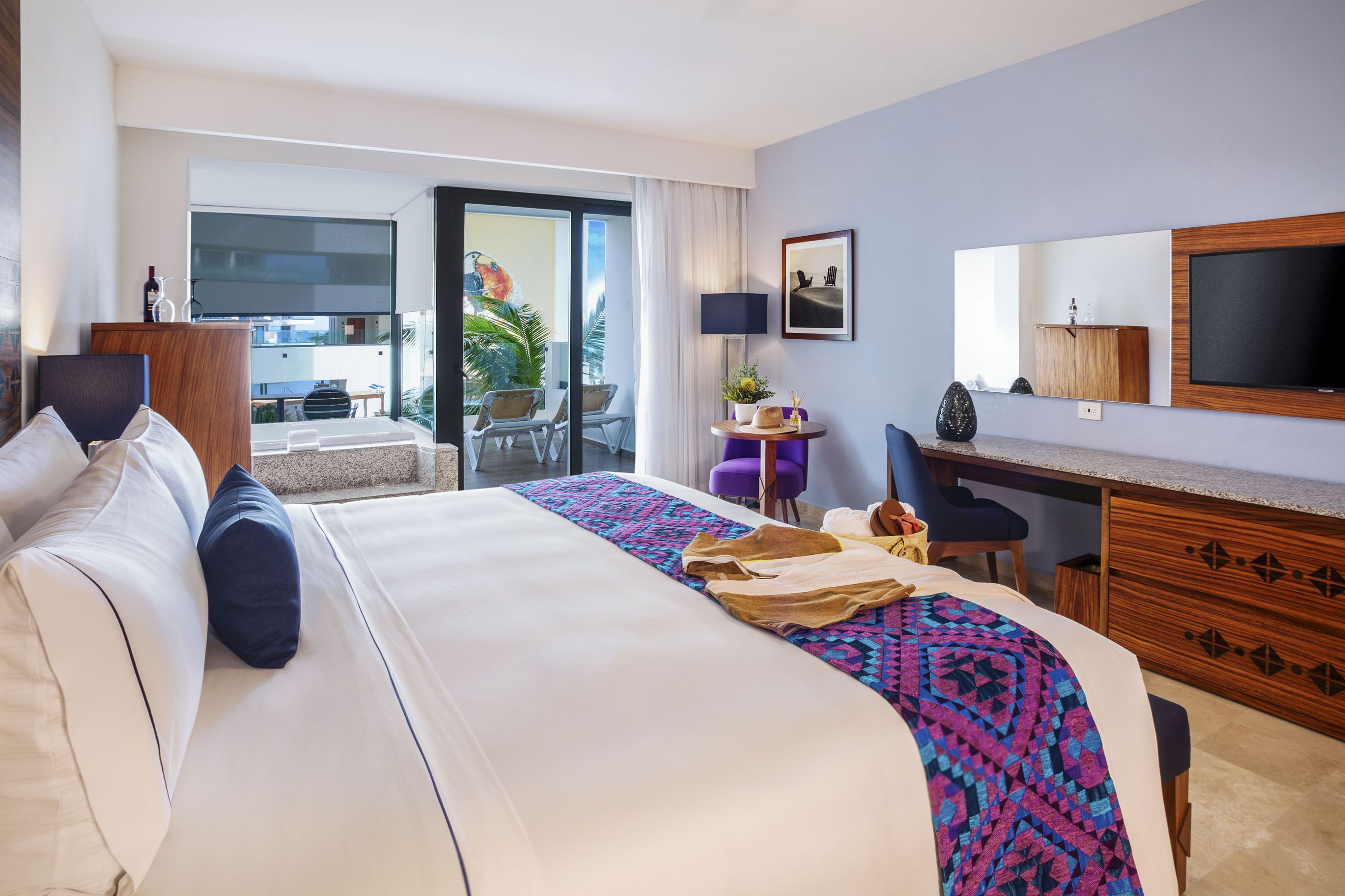 A hotel room with a large bed, a desk, a chair, a TV, and a balcony with a view.