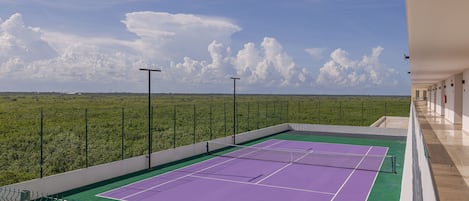 A tennis court with a net, surrounded by a fence, and a view of a green field under a clear sky.