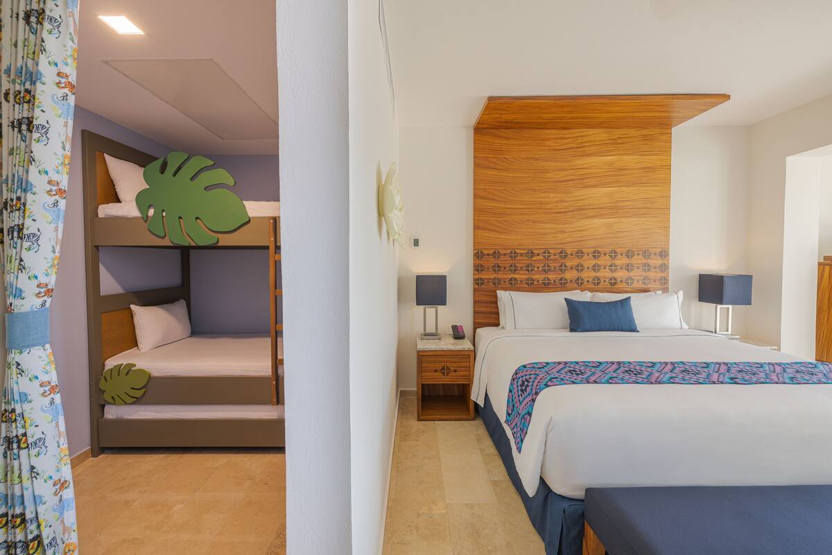 A hotel room with a bunk bed, a large bed, and a nightstand with a lamp.