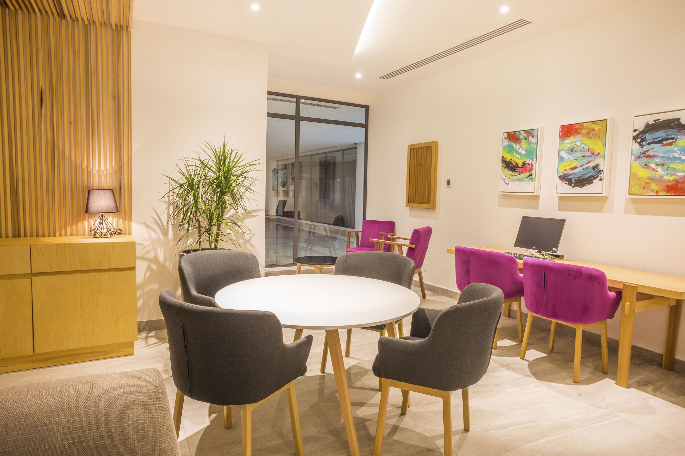A modern meeting room with a round table, grey and purple chairs, a wooden desk, and a laptop.