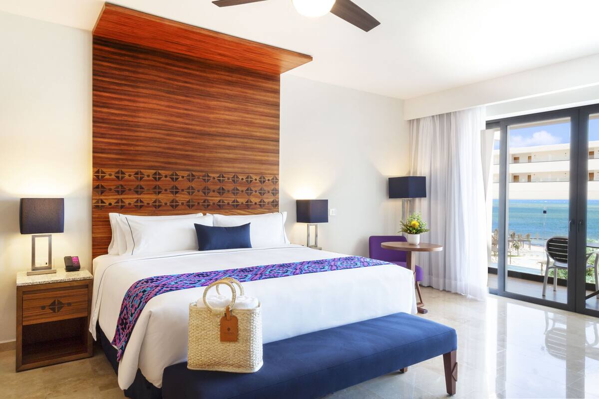 A hotel room with a large bed, a wooden headboard, a balcony with a view of the ocean, and a small table with a lamp.