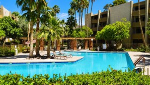 Ein Swimmingpool mit Liegestühlen, Palmen und einem mehrstöckigen Gebäude im Hintergrund.