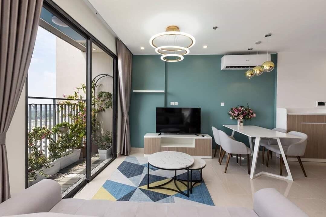 A modern living room with a TV, dining table, and chairs. There is a balcony with plants and a view of the sky.