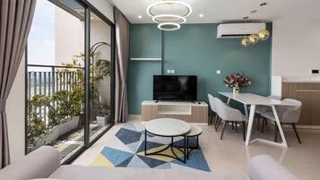 A modern living room with a TV, dining table, and chairs. There is a balcony with plants and a view of the sky.