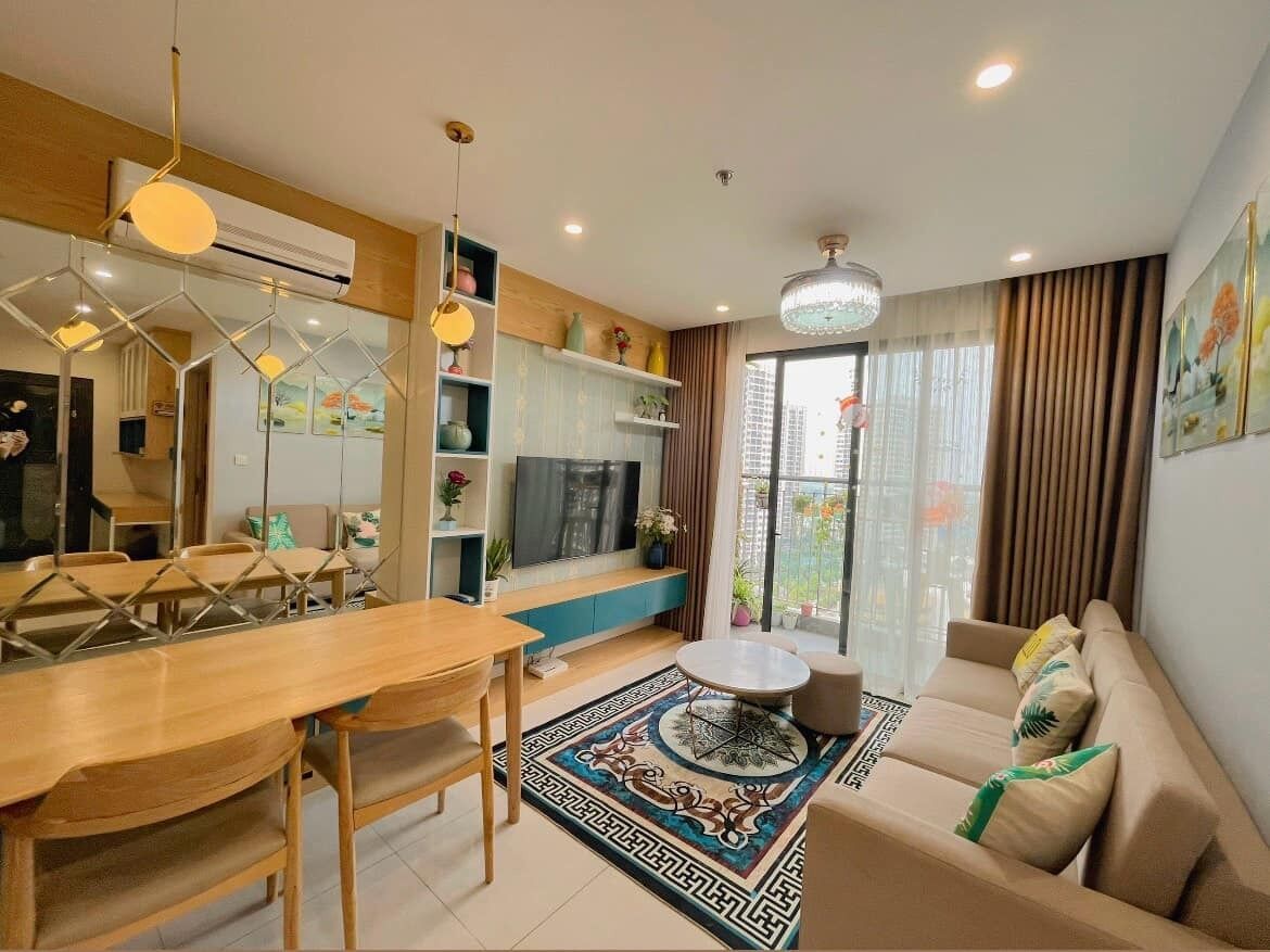 Ett modernt vardagsrum med en soffa, ett matbord och stolar. Det finns en TV som är monterad på väggen, en kristallkrona och ett stort fönster med gardiner.
