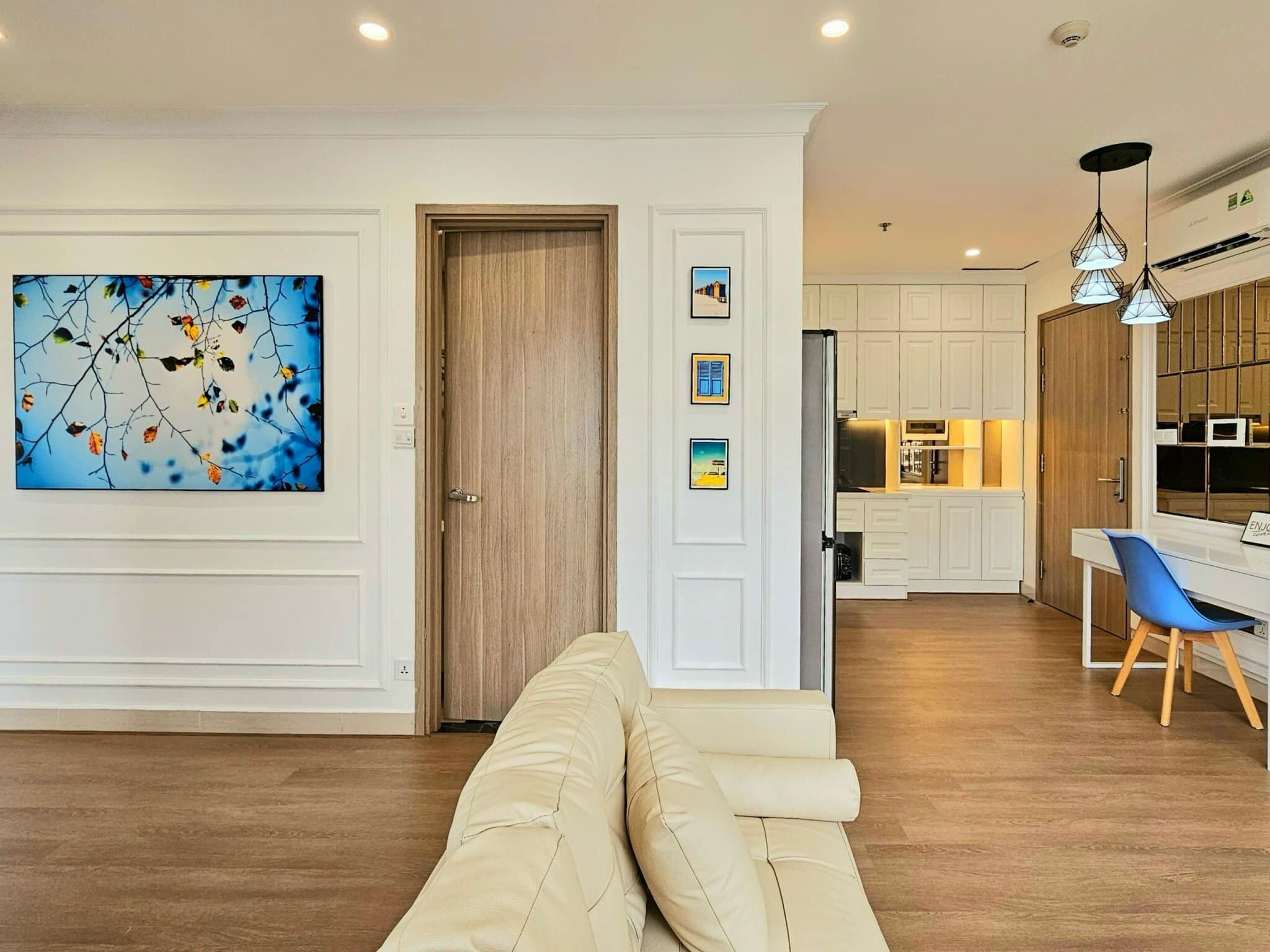 A modern living room with a beige sofa, a dining area with a blue chair, and a kitchen with white cabinets.
