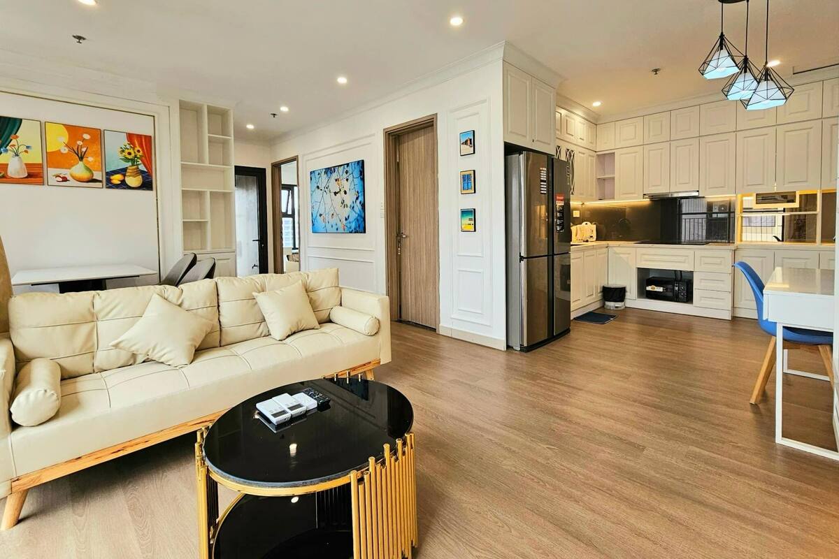 A modern living room with a beige sofa, a black coffee table, and a kitchen area with white cabinets and stainless steel appliances.