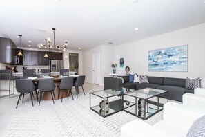 A modern living room with a glass coffee table, a grey sofa, and a dining area with a white table and chairs.
