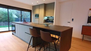 A modern kitchen with a large island, two bar stools, a built-in oven, and a view of the outdoors. - Luxueuse villa, piscine intérieure, plage à 700m (Saint Coulomb)