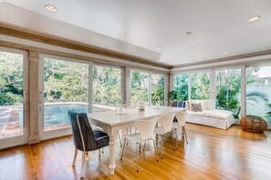 A spacious room with a large dining table, white chairs, a sofa, and a view of a swimming pool.
