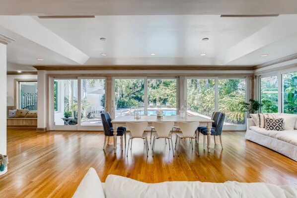 Un comedor amplio con una gran mesa y sillas, rodeado de ventanas que ofrecen vistas a una piscina y follaje.