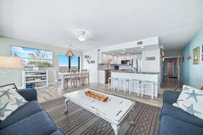 Un salon moderne avec une table basse blanche, des canapés bleus et un coin cuisine doté de meubles blancs et de tabourets de bar.