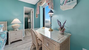 A bedroom with teal walls, a glass-top desk, wicker furniture, a lamp, and a vase with flowers. - Island Winds West 580 | Walk to HangOut! | Heated Pool! | Beach Getaways (Gulf Shores)