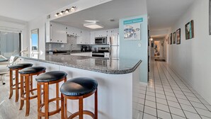 A modern kitchen with a central island, bar stools, and a view of the living area. - Island Winds West 580 | Walk to HangOut! | Heated Pool! | Beach Getaways (Gulf Shores)