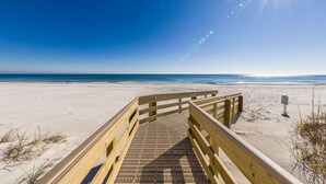 A wooden boardwalk leading to a sandy beach with a clear blue sky and sun shining. - Island Winds West 580 | Walk to HangOut! | Heated Pool! | Beach Getaways (Gulf Shores)
