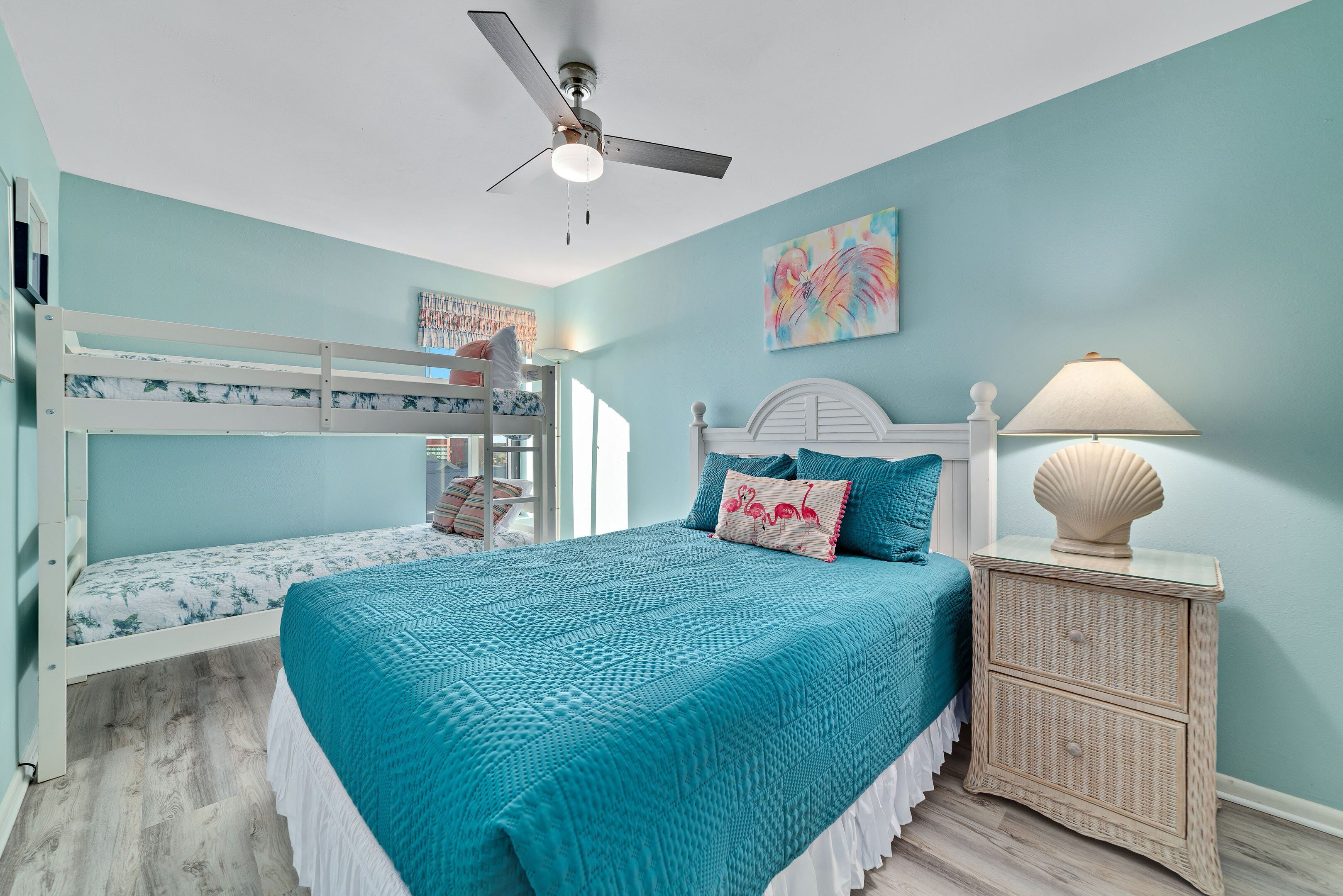 A bedroom with a bunk bed, a single bed, a ceiling fan, a nightstand with a lamp, and a wall painting.