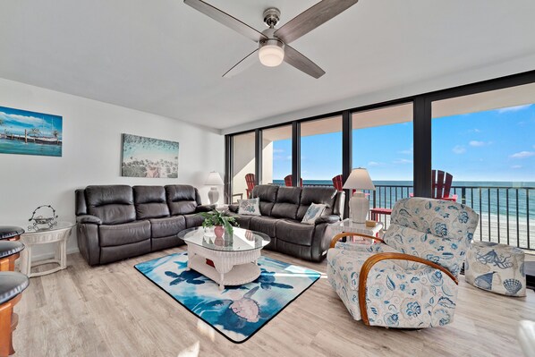 A spacious living room with a large window overlooking the ocean, a ceiling fan, and a modern fireplace. - Island Winds West 580 | Walk to HangOut! | Heated Pool! | Beach Getaways (Gulf Shores)