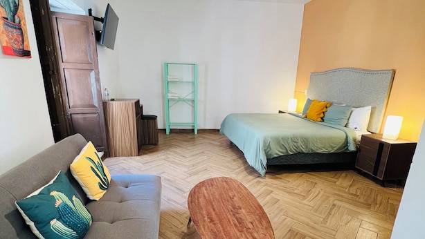 Une chambre moderne avec un canapé, une petite table ronde, un lit, un téléviseur fixé au mur et un parquet en bois disposé en chevrons.