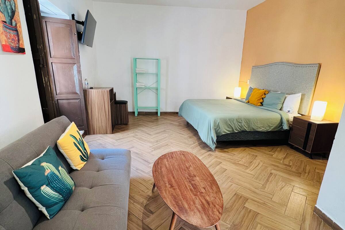 Een moderne kamer met een bank, een klein rond tafeltje, een bed, een aan de muur bevestigde televisie en een houten visgraatvloer.