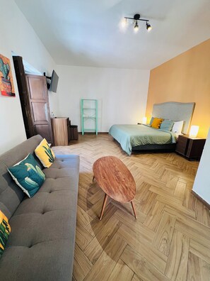 A modern room with a sofa, a small round table, a bed, a TV mounted on the wall, and a wooden herringbone floor.
