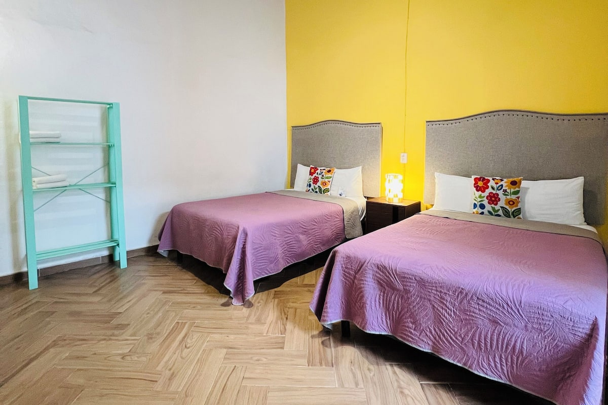 Een kamer met twee bedden, een houten visgraatvloer, een metalen rek en een gele wand.