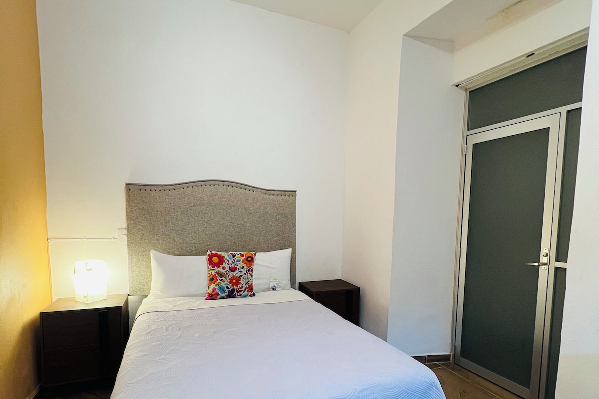 Een hotelkamer met een bed, nachtkastje, lamp, deur en een muur.