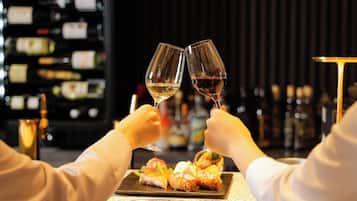 A bar setting with two people toasting with wine glasses, a plate of appetizers, and a background of wine bottles.