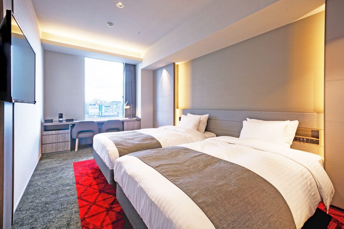 A hotel room with two beds, a desk, and a large window.