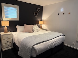 A bedroom with a bed, two bedside tables with lamps, a wall-mounted heater, and decorative heart-shaped wall art. - Snowman 3 Luxury stay Central Village & Parking. (Thredbo)