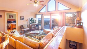 A spacious living room with a high ceiling, a large window with a view, a sofa, a bookshelf, and a ceiling fan.