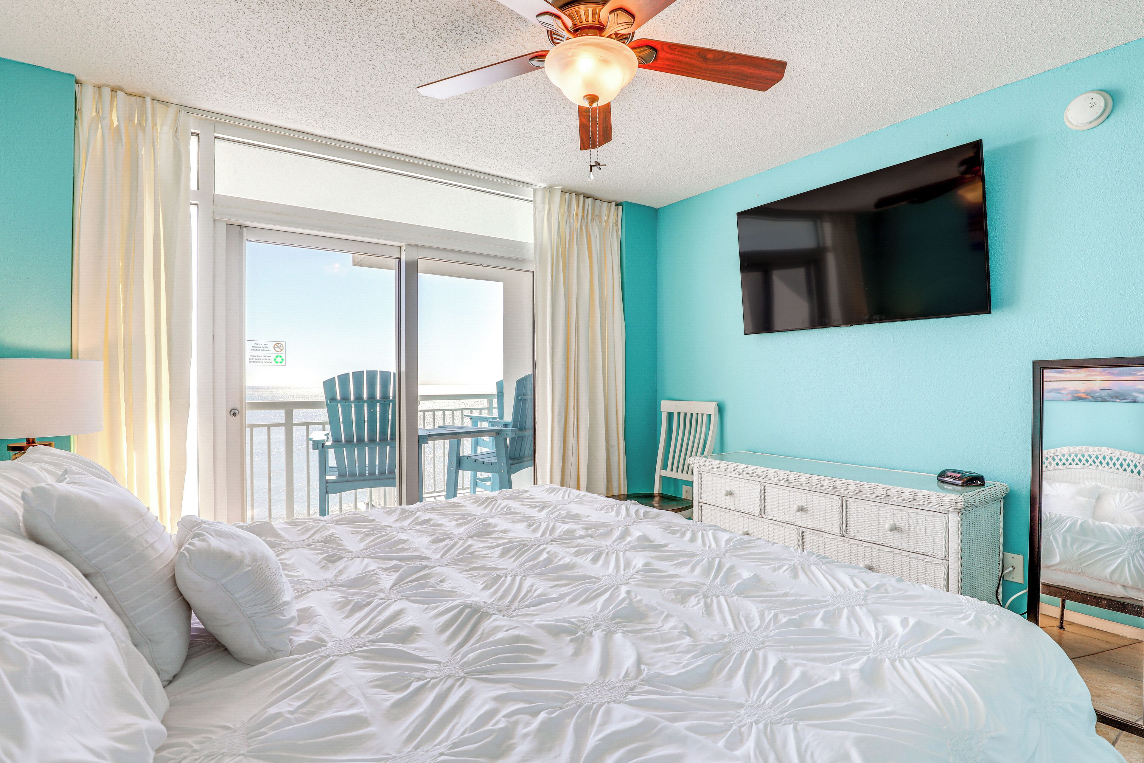 A bedroom with a large bed, a ceiling fan, a TV mounted on the wall, and a view of the outside.