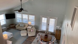 A spacious living room with a high ceiling, a ceiling fan, a round wooden table with chairs, a sofa set, and a television mounted on the wall.