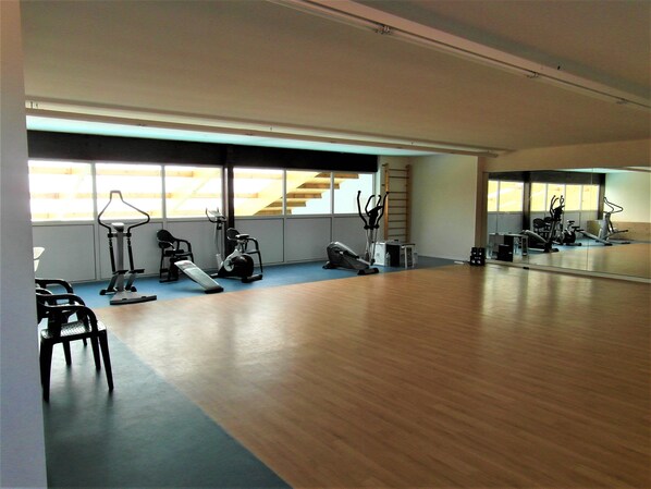Ein geräumiger Fitnessraum mit unterschiedlichen Trainingsgeräten, großen Fenstern und Holzboden.