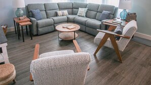 A modern living room with a sectional sofa, a round coffee table, and two armchairs. There is a ceiling fan, a lamp, and a small side table.